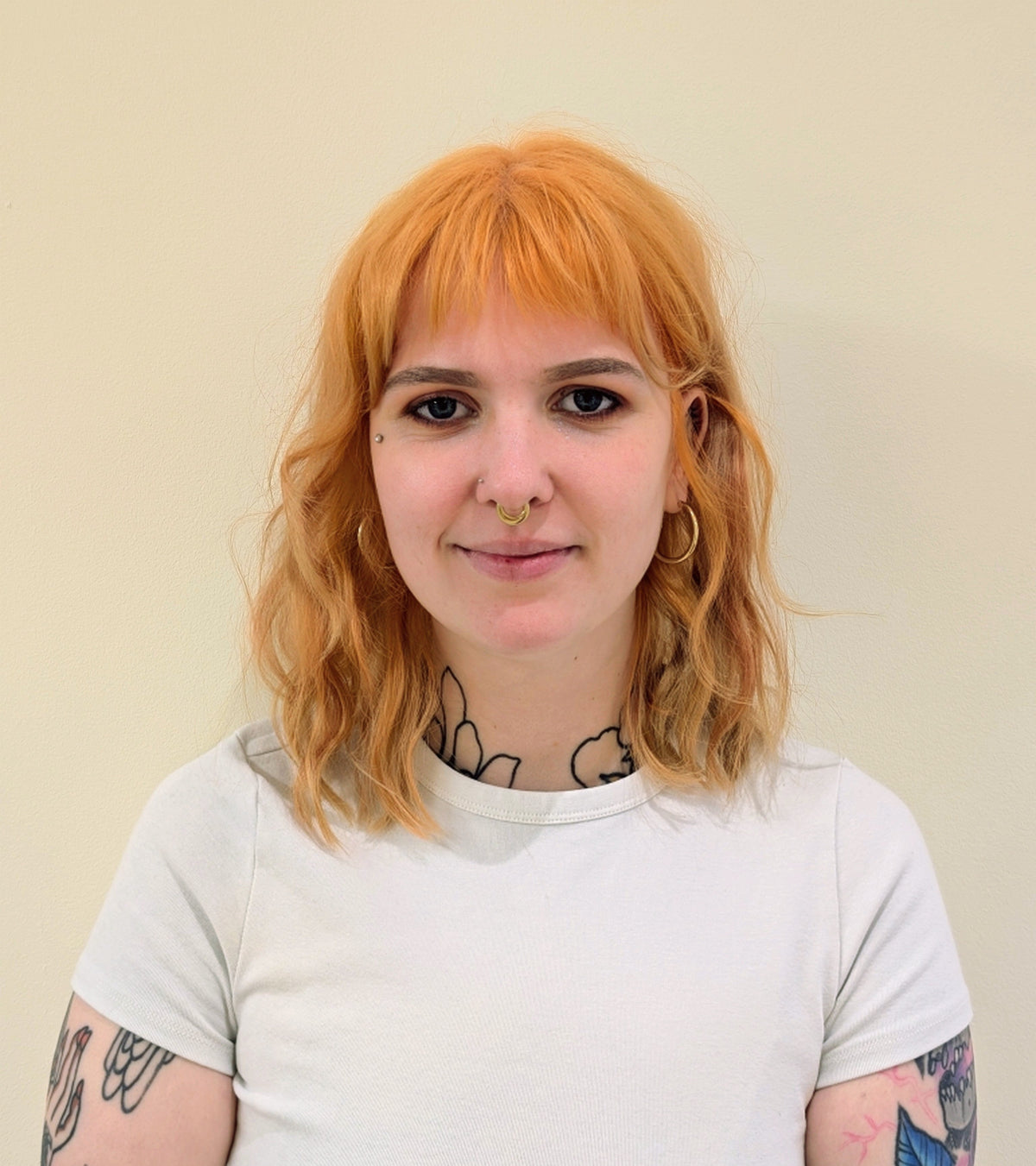 Person with red hair and tattoos wearing a white shirt against a plain background